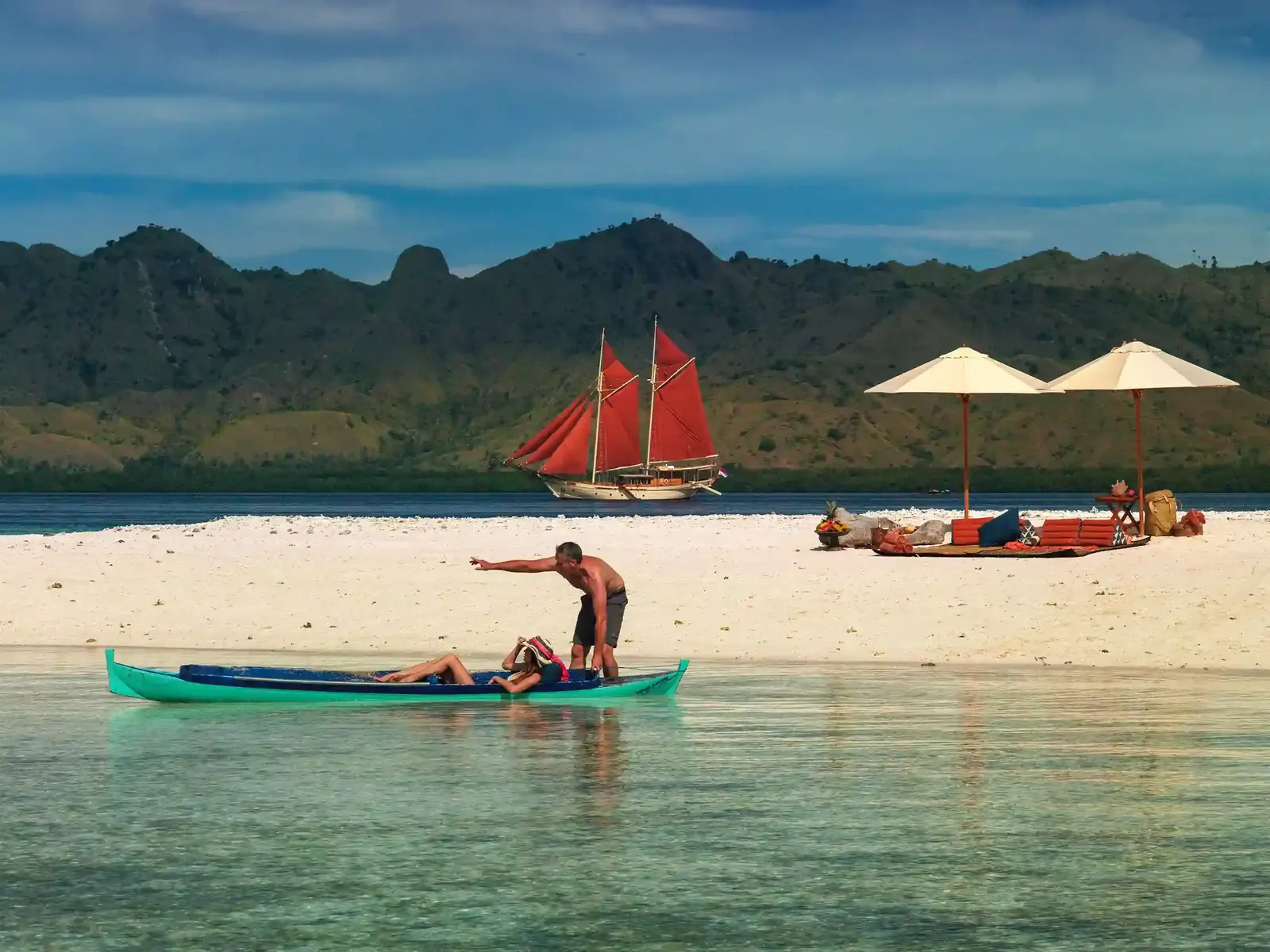 Guests relaxing on a Komodo sandbank, a red-sailed phinisi offshore